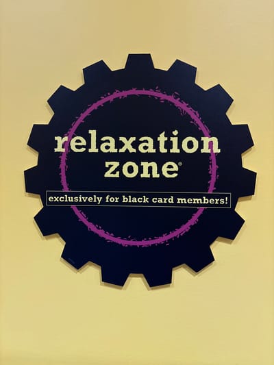 A dark blue gear-shaped sign with a jagged edge is mounted on a textured, pale yellow wall. Centered within the gear is a circular design, outlined in a distressed purple color. Inside the purple circle, the words "relaxation zone" are printed in white block letters, with "zone" appearing below "relaxation." The registered trademark symbol (®) is positioned to the right of "zone." Below the circular design, a white rectangle contains the text "exclusively for black card members!" in white block letters. The sign is illuminated from the front, casting a subtle shadow on the wall. No people are visible in the image. The location is the city of Tucson, United States of America.