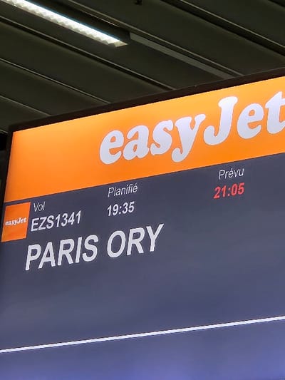 An illuminated digital display board, likely located within Geneva Airport (GVA) in Le Grand-Saconnex, Switzerland, occupies the majority of the frame. The upper section of the board features an orange background with the white "easyJet" logo prominently centered. Below this, against a dark gray background, white text details flight information. The left column shows a smaller "easyJet" logo followed by "Vol" and the flight number "EZS1341," indicating a flight to "PARIS ORY" (Paris Orly). The middle column displays "Planifié" (Scheduled) with a time of "19:35." The right column shows "Prévu" (Estimated) with the updated time "21:05" rendered in red text. Above the display board, parts of a dark gray ceiling structure are visible, featuring recessed, linear fluorescent light fixtures with a distinct textured or corrugated diffuser pattern. No people, specific actions, or interactions are visible in the scene. The environment is consistent with an indoor airport terminal setting.