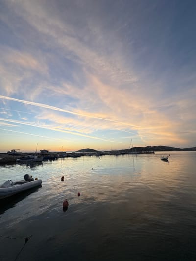 A tranquil waterscape during a sunset. The sky features streaky clouds with soft yellow hues. A coastline with docked boats and buildings runs along the horizon. The water's surface reflects the sky's colors. Red buoys float. A small island or hill is visible in the distance. Vapor trails cut through the sky.