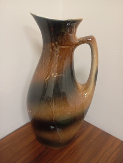The image shows a tall, ceramic pitcher with a glossy, mottled glaze that transitions from dark brown and black at the bottom to lighter brown and beige towards the top. The pitcher has a distinctive handle and a flared lip. It is positioned on a wooden surface, likely a table or shelf, with a plain, light-colored wall in the background. The lighting appears to be artificial, casting soft shadows. There are no people or visible text in the image. The scene is static, with the pitcher being the sole focus. The overall impression is one of a decorative or functional ceramic object. The provided location context of Ararat, Armenia, does not offer specific visual cues within the image itself.