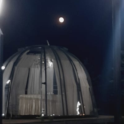 The image depicts a geodesic dome-shaped structure at night, with a full moon visible in the dark sky above. The dome is illuminated from within, revealing draped white fabric and what appears to be a table covered with a white cloth. The exterior of the dome is constructed of dark, segmented panels. The setting appears to be outdoors, possibly in the city of Sosnovka, Kyrgyzstan, given the request. The overall scene suggests a nighttime event or venue within a dome structure, under the moonlight.