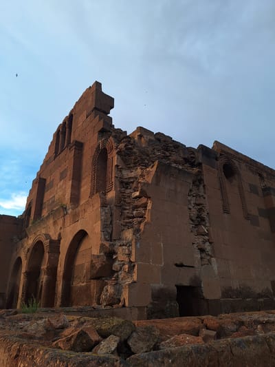 The image shows a portion of a damaged stone building, possibly a church or historical structure, located in Anipemza, Armenia. The architecture features brick or stone construction with arched windows or openings. There is visible deterioration and collapse of the structure, with fragmented walls and debris. The sky is partly visible in the background.