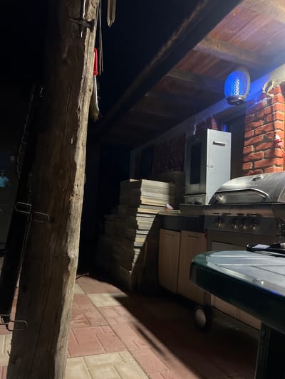 This image depicts a covered outdoor area at night in Debelt, Bulgaria. A large, rough-hewn wooden post occupies the left foreground, displaying a metallic hammer and other tools hanging from its surface, along with a red-handled item. To the left of the post, a dark, ladder-like structure is partially visible. The ground is paved with geometric tiles in alternating light and dark reddish-brown hues, extending towards the background. In the midground, a substantial stack of rectangular, light-colored paving stones or tiles is positioned. An outdoor cooking station is centrally located, featuring a silver gas grill with a closed lid and control knobs. Adjacent to the grill on the left, a light-colored wooden cabinet unit with two doors is visible, next to a tall, grey metallic unit, possibly a smoker or storage cabinet, positioned against a brick wall. Overhead, a ceiling constructed from wooden beams and planks supports a prominent oval-shaped blue light fixture, likely an insect zapper, emitting a distinct blue glow. Sections of a red brick wall are visible in the background, behind the cooking station. Artificial light sources create defined shadows across the paved surface and objects. No individuals are present in the scene.