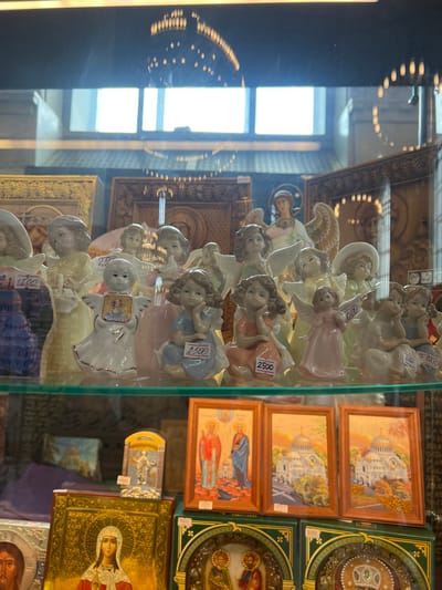 The image displays a close-up view of a shop window filled with religious figurines and icons. In the foreground, several ceramic figurines of angels are arranged on a glass shelf. These angels vary in size and pose, some holding books or baskets, others in seated positions. Price tags are visible on some of the figurines, with denominations such as "2500" and "1500" in Russian currency.

Behind the angel figurines, a larger ceramic figure of an angel with outstretched wings is visible. The background of the shop window features a display of framed religious icons. One prominent icon depicts the Virgin Mary, richly adorned with gold leaf and intricate patterns. Other icons show depictions of saints and architectural landmarks, possibly churches or cathedrals, with stylized domes.

The overall environment suggests a religious goods store. The lighting in the scene is a mix of artificial light from within the shop and natural light filtering in from a window or skylight in the background, which is slightly obscured by reflections. The presence of icons depicting what appear to be Russian Orthodox churches, combined with the specific mention of Kronstadt, Russia, suggests this store might be located in Kronstadt.