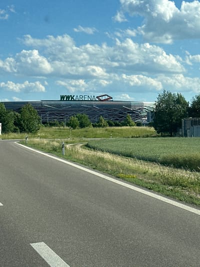 The image shows a highway leading to the WWK Arena, a football stadium located in Augsburg, Germany. The stadium is a large, modern structure with a distinctive facade. The sky is blue with white clouds, and the surrounding area is green with trees and fields. The road is paved and has white lines marking the lanes. There are no people visible in the image.