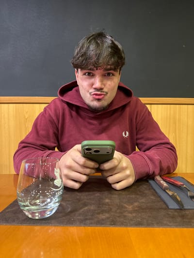 A young adult male is seated at a light-brown wooden table in an indoor dining establishment, likely a restaurant, within the city of Peso da Régua, Portugal. The exact location within the city is not identifiable from the visual information. The male, with dark, wavy hair, a slight mustache, and a goatee, looks directly at the camera with a pursed-lip expression. He wears a maroon hooded sweatshirt featuring a white embroidered laurel wreath logo on the left chest. His hands hold a dark green-cased smartphone horizontally, with thumbs positioned over the screen. On the dark brown placemat in front of him, to his left, is a clear, stemless glass containing a clear liquid and an etched design. To his right, a set of cutlery (knife and fork with reddish-brown wooden handles) rests on a dark napkin. The background consists of light-brown wooden paneling at mid-height, topped by a dark grey or black wall surface.