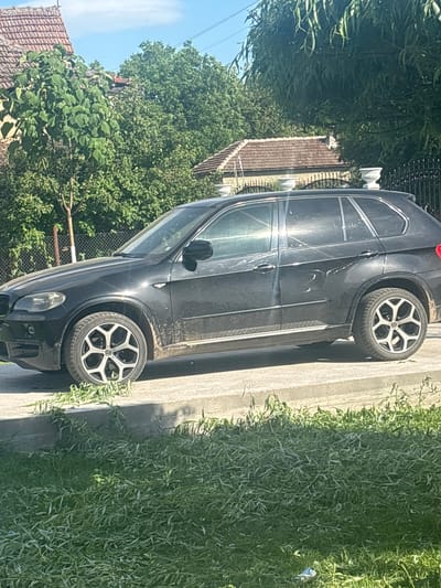 A black BMW SUV is parked on a concrete curb in Septemvritsi, Bulgaria. The car faces left, with its front end partially obscured by green vegetation. Its wheels feature a silver and black multi-spoke design. The exterior of the vehicle appears dusty. In the background are trees, a fence, and a building with a red-tiled roof. A bright sky is visible above the trees.
