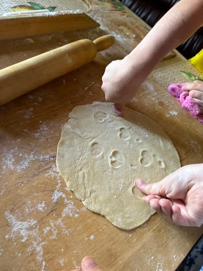 This image depicts a close-up overhead view of two pairs of hands interacting with dough on a wooden surface. The scene is not located in Valday, Russia. One person's hands are gently pressing indentations into a large, circular piece of light-colored dough that is spread across the wooden table. These indentations appear to be in the shape of small hearts. Another person's hands are visible on the right side, holding a small, pink, textured cloth. A wooden rolling pin lies horizontally in the upper left quadrant of the image, and another similar object is partially visible behind it. The wooden surface is dusted with flour, indicating food preparation. The lighting is warm and natural, casting soft shadows. The background is out of focus but suggests an indoor setting with dark furniture.