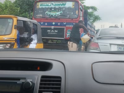 The image depicts a traffic scene in Iju, Nigeria. In the foreground, the interior of a car is visible, showing a dashboard with a radio display and a mode knob. In the middle ground, a yellow auto-rickshaw is parked, with two people inside wearing white attire. To the right of the auto-rickshaw, a red and blue truck is present. A man, wearing a black t-shirt and light-colored shorts, is standing next to the truck, holding a yellow jerrycan and appearing to pour liquid into the silver car behind the truck. The silver car is a Honda Accord with a license plate that reads "LAGOS SMK-49". The background shows lush green trees and overcast skies. The overall impression is one of a busy and potentially congested street.