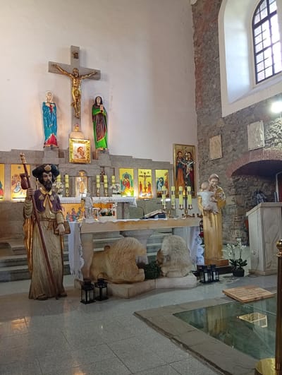 The image depicts the interior of a church in Sobótka, Poland. The central focus is an altar area with several religious statues and paintings. In the foreground, a life-sized statue of a bearded man in pilgrim attire stands to the left, holding a staff and a book. To the right of the altar, a statue of Saint Anthony holding the infant Jesus is prominent. The altar itself is covered with a white cloth and decorated with candles and floral arrangements. Behind the altar, a large crucifix with a gilded figure of Jesus is mounted on the wall, flanked by statues of the Virgin Mary and another female figure. Various religious paintings are displayed on the walls, some depicting biblical scenes. The church interior features stone and plastered walls, and a large arched window with multiple panes is visible on the right side. The floor is tiled, and a section with a glass covering reveals an area below. Two stone lion sculptures are positioned in front of the altar.