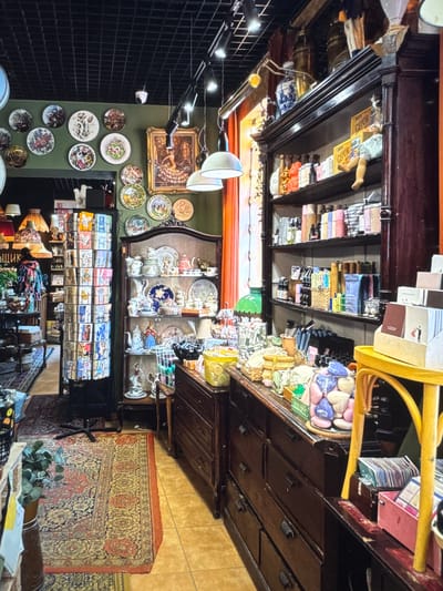 The image depicts an interior view of a shop, likely an antique or curio store, located in Novinki, Russia. The shop is filled with a variety of decorative and functional items. Dominating the right side of the frame is a tall, dark wooden cabinet with multiple shelves displaying an assortment of merchandise. These items include bottles, boxes, and decorative objects. Below the shelves, a dark wooden dresser with several drawers is visible, topped with more displayed items such as baskets, jars filled with colorful objects, and small boxes.
On the left side of the image, a green wall is adorned with numerous decorative plates. In the center, a tall rotating display rack filled with postcards or small booklets is positioned. Further to the left, antique lamps with patterned shades are visible, along with other display shelves and furniture. The floor is covered with Persian-style rugs. The overall atmosphere suggests a cluttered yet curated retail environment, common in shops selling vintage or artisanal goods. The lighting is a mix of overhead fixtures and display lighting, creating a warm and inviting ambiance. There are no visible people actively engaged in any action within the frame.
