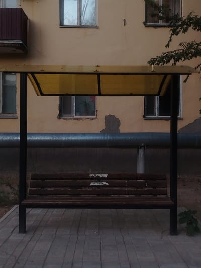 This media file depicts an outdoor scene in Baikonur, Kazakhstan, featuring a simple wooden bench with a yellow canopy, positioned on a paved walkway. The bench is dark brown and weathered, showing signs of age and use. It is supported by black metal legs. Above the bench, a translucent yellow canopy offers protection from the elements, attached to black metal framing.
Behind the bench and canopy, a light yellow building with multiple windows is visible. Some windows are intact, while others appear damaged or boarded up. A large, silver pipe runs horizontally across the middle of the building's facade. The building's exterior shows signs of wear, with peeling paint and some discoloration. A balcony with a dark red railing juts out from the left side of the building. To the right, a leafy branch hangs into the frame from a tree, its dark green foliage contrasting with the building's warm tones.
The ground in front of the bench is paved with rectangular gray tiles, laid in a regular pattern. To the sides of the bench, sparse vegetation and dirt are visible. The overall lighting suggests it is either early morning or late evening, with soft, diffused light casting subtle shadows. There is no discernible activity or event taking place; the scene is static and quiet, conveying a sense of stillness and perhaps abandonment. No text is visible in the image.