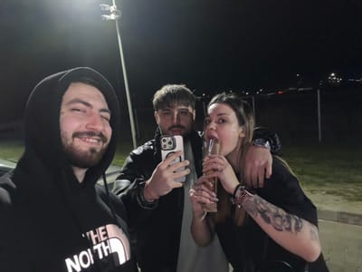 A night-time image captures three individuals outdoors in Frătești, Romania. On the left, a male with a beard and dark hooded sweatshirt featuring "THE NORTH FACE" logo is smiling and winking, seemingly taking a selfie. In the center, a second male with a mustache holds a silver-colored smartphone with a multi-lens camera system in his right hand. He has his left arm draped over the shoulder of the female on the right. This female, with dark hair pulled back, displays a heavily tattooed left arm, wears a watch, multiple bracelets, and a dark short-sleeved top. She is depicted drinking from a tall, clear glass or bottle, with her head tilted back and mouth open. The background is dark, illuminated by a prominent pole-mounted light fixture in the upper left of the frame. Distant lights and faint structures are visible further back, indicating an open, illuminated outdoor area, possibly a parking lot or roadside location.