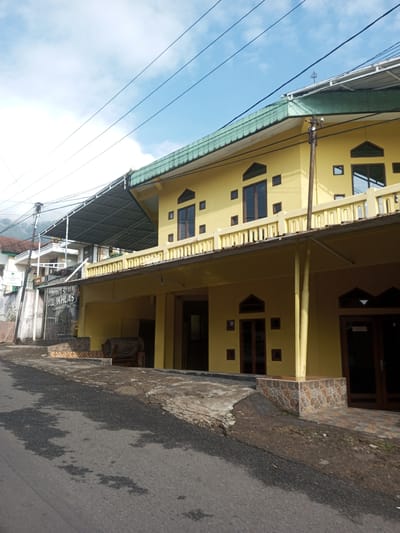 The image shows a two-story yellow building in Mya Sit Kaing Ywar Thit, Myanmar. The building has a green roof and a balcony with a balustrade. There are windows and doors with dark frames on both floors. A canopy extends over the ground floor entrance. A section of the street is visible in the foreground. Power lines are strung across the top of the frame. A portion of another building is visible to the left of the yellow building.