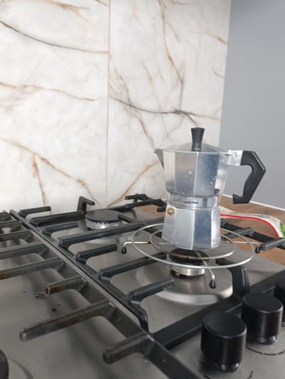 A stovetop espresso maker, also known as a moka pot, sits on a gas stove. The moka pot is made of silver-colored metal and has a black handle and knob. It is placed on a circular metal trivet that is resting on the grates of the stove. The stove is made of stainless steel and has four burners, two of which are visible in the foreground. The background shows a tiled wall with a marble-like pattern and a grey wall. The scene appears to be a kitchen. The location is not identifiable as Marshyntsi, Ukraine.