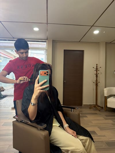 A man in a red GUESS t-shirt is cutting a woman's hair in what appears to be a salon. The woman is seated in a brown chair, covered with a black protective cape, and holding a green smartphone up, possibly taking a picture or recording the process. The salon has a modern design, with light-colored walls, a dark wooden door, and a coat rack on the wall. The location could be in Villahermosa, Mexico.