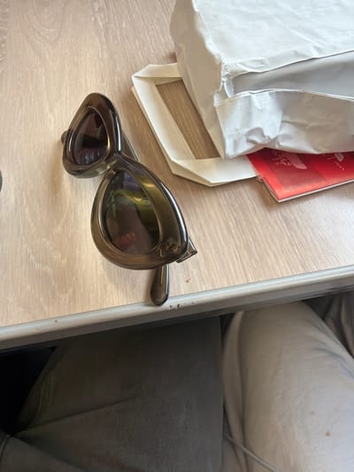 A still life scene, possibly indoors, depicts a light-colored wooden-like surface, possibly a table. On the left side, there's a white paper bag, partially open, and a red-colored rectangular object with what looks like printed designs. A pair of dark-framed sunglasses with thick rims and oval-shaped lenses is situated towards the center-right. To the right of the frame is a portion of a grey upholstered surface, likely part of a seat.

Without additional visual context, it's not possible to determine a specific location in Bassetlaw, United Kingdom.