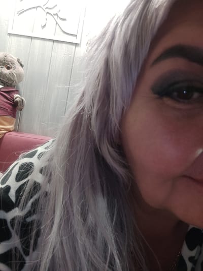 A close-up photograph shows a woman with long, light purple hair partially obscuring her face. Her right eye is visible, with dark eyeshadow and defined eyebrow. A portion of her cheek and mouth are also visible. She is wearing a black and white patterned top. In the background, out of focus, is a stuffed animal seated on a red surface. Behind the stuffed animal is a white wall with a white decorative cutout. The image does not provide enough information to determine the location in the city of Kotlas, Russia.