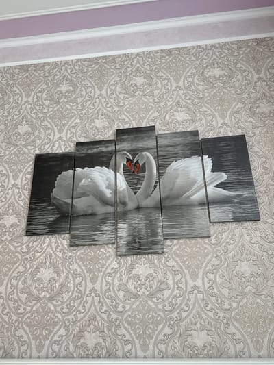 The image shows a wall in a room. The wall is covered with light-colored, damask-patterned wallpaper. A five-panel canvas print of two swans forming a heart shape with their necks is mounted on the wallpaper. The swans are white and are depicted in black and white, with their orange beaks being the only touch of color. The print is centered on the wall. Above the wallpaper, there is white crown molding, and above that, a section of a wall painted light purple. The overall scene suggests a domestic interior, likely a living room or bedroom. There are no people visible in the image.