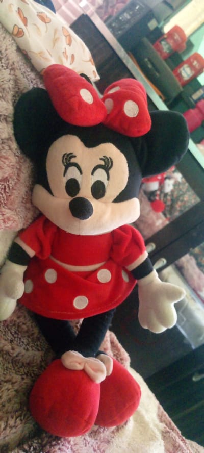 A plush Minnie Mouse toy is angled diagonally from top left to bottom right, resting on a textured surface. The toy is primarily black and red with white polka dots on its dress and shoes. It has large, round ears, a pale yellow face with black eyes and eyelashes, and a dark blue nose. A red bow with white polka dots adorns its head. The toy wears a red dress with white polka dots and a white waistband. Its feet are covered in red shoes with white polka dots and a pink bow detail. In the background, a blurred reflection in what appears to be a glass surface shows shelves with various items, including what look like red bottles and other toys. The environment is indoors and appears to be a domestic setting. The location is specified as Marhamat, Uzbekistan.