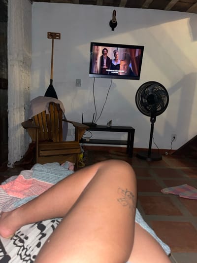A tilted room features a wall-mounted television playing a show, a black standing fan, and a wooden chair. A wine bottle holder is mounted high on the wall. On the right side, a person's legs with a tattoo are visible, resting on a blanket with a black and white pattern. The foreground is blurry, while the background objects are relatively clear. The room has a rustic feel with brick walls.