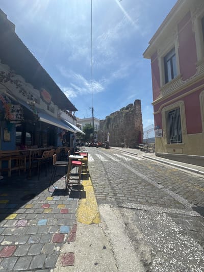 The image captures a sunlit street scene in Kavala, Greece, likely in the old town near the fortifications. On the left, a blue-fronted building with a dark awning houses an outdoor cafe. Tables and chairs, some with red seats, are set up along the sidewalk, which is paved with colorful, square-shaped tiles. The building's facade is adorned with floral decorations and signage.
The street itself is paved with cobblestones, with a zebra crossing visible further up. To the right of the street, a large, ancient stone wall, part of the city's fortifications, stands prominently. Adjacent to the wall, a light-colored building with ornate decorative elements and barred windows is partially visible. Further in the background, more buildings and a power pole are seen under a bright blue sky with scattered clouds. A few motorcycles and scooters are parked along the street. The sun is high, casting distinct shadows.