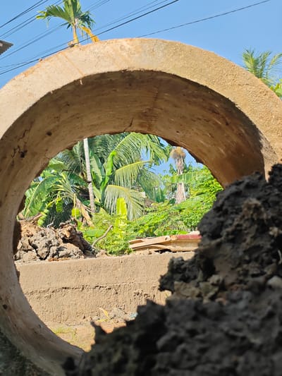 An eye-level, medium shot captures a concrete culvert in Ubitpayaitek, Indonesia, framing a vibrant tropical landscape under a clear blue sky. The foreground is dominated by the rough, textured surface of the concrete culvert, appearing aged and weathered with visible cracks and a sandy hue. To the right, a pile of dark, clumpy soil obscures a portion of the view.
Through the circular opening of the culvert, a lush scene unfolds. Dense, green foliage, characteristic of a tropical environment, fills the midground. Prominent banana trees with their large, broad leaves are visible, along with palm trees, their fronds reaching towards the sky. Interspersed are smaller, leafy green bushes and plants, creating a rich tapestry of vegetation. In the background, a faint hint of buildings with red roofing can be seen amongst the trees, suggesting a rural or semi-urban setting.
The lighting suggests a bright, sunny day, casting clear shadows and highlighting the vibrant greens of the vegetation. The overall impression is one of a construction site or a natural area undergoing development, with the culvert acting as a natural framing device for the picturesque tropical scenery beyond. There are no people or animals visible in the frame.