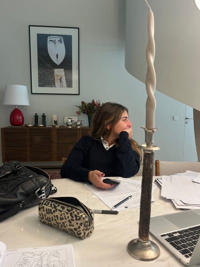 An adult female is seated at a light-colored table in an interior space within Lordelo do Ouro e Massarelos, Portugal. The woman, with long brown hair, wears a dark blue V-neck sweater over a white collared shirt. Her head is turned to the right, and her right hand rests on her chin. Her left hand holds a dark-colored smartphone. On the table to her left, a black leather bag and an open leopard-print pencil case are present. Directly in front of her, white papers with text and diagrams, along with a black pen and marker, are visible. A silver-colored laptop is partially visible to her right. In the foreground, a tall, dark-colored candlestick with a silver base and top holds a light-grey twisted candle. The background features a light-colored wall with a large framed black-and-white print depicting a boat and people on water and a pier. Beneath the print, a wooden slatted sideboard holds a red ceramic lamp with a white shade, two green decorative objects, a white sculpture, a silver tea set, and a vase of flowers. To the far right, a segment of a white modern staircase and a light-colored door with a metallic handle are visible.