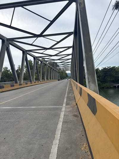 The image shows a view of a bridge spanning a body of water in Lamkruet, Indonesia. The bridge is constructed with a steel truss superstructure and a concrete deck. The road on the bridge has a yellow and black barrier on the right side, with a white lane divider. Trees and vegetation line the banks of the water. The sky is overcast. On the bridge, there are a few motorcycles and a person on foot visible in the distance. The scene appears to be during daylight hours.