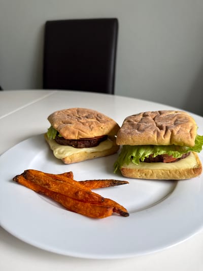 A close-up, eye-level photograph captures a meal consisting of two small burgers and a serving of sweet potato fries presented on a white, round ceramic plate. The scene is situated in Hazlemere, United Kingdom. The plate occupies the majority of the lower half of the frame.

The burger on the right, positioned centrally, features a square, lightly browned, textured bun encasing a dark brown patty, a melted slice of light yellow cheese, a red tomato slice, and a visible layer of bright green lettuce. The burger on the left, slightly smaller and positioned behind the right one, uses a similar bun, containing a dark patty and a more visibly melted, flowing slice of light yellow cheese, with other fillings less distinct.

To the lower-left of the plate, several elongated, vibrant orange sweet potato fries are arranged, some exhibiting darker, crispier edges. The white surface underneath the plate is a table or countertop, reflecting ambient light. In the blurred background, a plain light grey wall is visible, with a dark brown, upright chair back positioned towards the center-right. No individuals are present in the image.