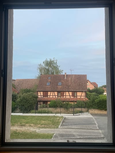 The image shows a view through a window of a half-timbered house in Gerstheim, France. The house has a red tile roof and cream-colored walls with dark brown timber framing. There are two dormer windows on the roof and shutters on the windows of the house. In front of the house, there is a green lawn and a paved area. The sky is a light blue with some clouds. The overall scene is peaceful and serene, typical of a small French village.