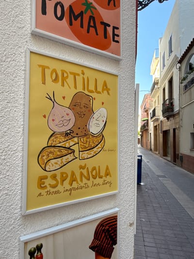 The image shows a close-up of several framed artworks displayed on a textured white wall. The artwork in the foreground is a yellow poster with a drawing of a potato, an onion, and an egg, all with cartoon faces. The text on the poster reads "TORTILLA ESPAÑOLA" and "a three ingredients love story". The artwork above this one is partially visible and appears to be a poster with the word "TOMATE" and a tomato graphic.
The wall the art is displayed on has a rough, stucco-like texture. To the right of the wall, a narrow cobblestone street is visible, lined with buildings with balconies. The sky above is a clear blue. The overall scene suggests an outdoor display of art, possibly in a commercial area or a street market in Tossa de Mar, Spain.
