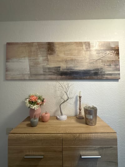 A wooden dresser with two drawers is positioned against a white, textured wall. Above the dresser, a long, horizontal abstract painting is displayed. On the dresser, from left to right, are a grey apple figurine, a pink apple figurine, a pink vase filled with a bouquet of white and pink flowers, a silver decorative tree, a pink candle holder with a single white candle, and a silver metallic container with a bow and label, likely a candle or potpourri. The dresser has silver handles on its drawers. The scene appears to be an indoor domestic setting.