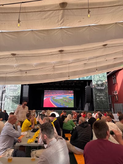 The image depicts an outdoor gathering under a large, draped canopy, likely a temporary structure for an event. In the background, a large screen displays a sports match, possibly football, with a stadium crowd visible. To the left and right of the screen are large banners. The left banner has the text "FOYN- HAGEN" and "Summer Racing" in white lettering against a green background with an image of a tree and leaves. Below this are smaller logos for "4human", "NORLIOG", "MENY", "DNB" and "COFFICO". The right banner is identical. To the right of the banners and screen is a red wooden building with white window frames.

In the foreground and midground, numerous people are seated at long tables, some with benches. They appear to be watching the screen or interacting with each other. Many are holding drinks, and some have food in front of them. The atmosphere seems relaxed and social. Several individuals are wearing yellow shirts.

The overall scene suggests a public viewing event for a sporting competition, possibly at a venue in or near Tønsberg, Norway, given the presence of the "FOYN- HAGEN" banner which appears to be a local establishment. The lighting suggests it is daytime, though the canopy creates a shaded environment. There are hanging light bulbs visible, indicating the potential for evening use as well.