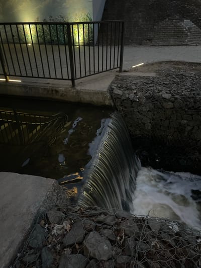 The image shows a concrete canal with water flowing over a weir. The canal is lined with stone and wire mesh on the sides. A metal railing runs along the side of the canal. The water is flowing rapidly, creating a small waterfall. The reflection of the railing can be seen in the water. The area is illuminated by artificial lighting, visible as bright spots in the water and along the walkway. The location is likely Chernyakhovsk, Russia, as inferred from the user query. The scene appears to be a man-made waterway feature within an urban environment.
