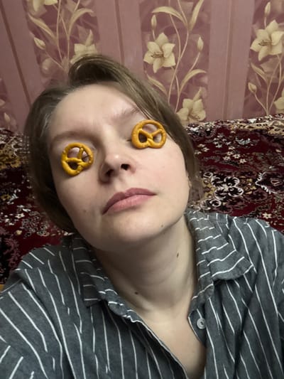 An indoor, close-up shot captures a person, likely a young adult woman, lying down with a playful expression. Two small, golden-brown pretzel snacks are placed over her closed eyelids. She is wearing a grey shirt with thin white horizontal stripes. Her light brown hair frames her face, and her lips are slightly parted.

The background features two different patterned fabrics. To the left, a light-colored fabric with a subtle floral design is visible, possibly curtains or wallpaper. To the right, a darker, richly patterned fabric with red, gold, and black motifs, resembling an oriental rug or blanket, forms the backdrop. The lighting is even, suggesting an indoor setting without specific time-of-day indicators. The overall mood of the image is lighthearted and humorous. No text is visible in the image. The scene takes place in Aksay, Kazakhstan.