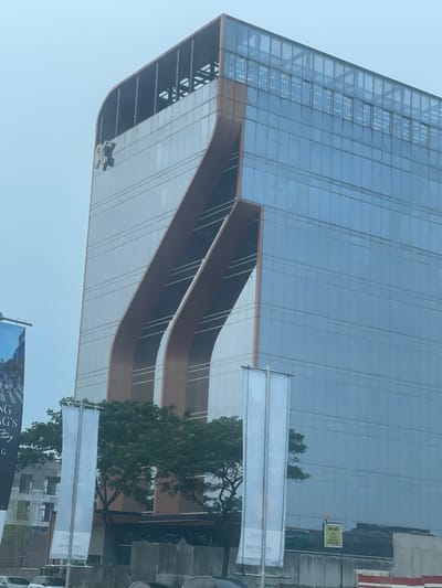 The image shows a modern, multi-story building with a glass facade and distinctive curved architectural elements. A silver logo is visible near the top of the building. In the foreground, there are two vertical banners, one partially obscured by a tree. A street sign with the text "HATI-HATI" (Indonesian for "Be careful") and a warning symbol is also visible. The sky is overcast. The building is identified as the Menara Astra in Jakarta, Indonesia, not Medan.