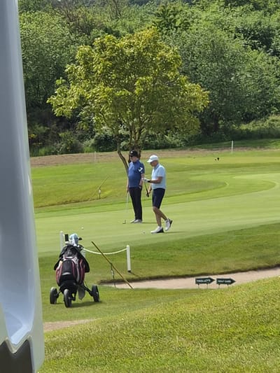 The image depicts two individuals playing golf on a green golf course. One person is wearing a blue shirt and black pants, while the other wears a white hat, light blue shirt, and dark shorts. Both appear to be actively engaged in the game. A golf bag is visible to the right of the players. A sign indicates "Trolleys" and "Golf Cars". The background includes trees and grass. The location is Lenningen, Luxembourg.