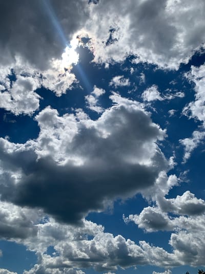 The image captures a dramatic sky filled with a mix of dark and light clouds against a deep blue sky. The sun is partially obscured by clouds, creating a bright lens flare and casting rays of light through the atmospheric haze. Some clouds are dense and grey, suggesting rain or storm activity, while others are bright white and fluffy, illuminated by the sun. The perspective is from below, looking upwards, emphasizing the vastness of the sky. The scene appears to be an outdoor environment, likely in Pokotylivka, Ukraine, given the context. No people or specific structures are visible.