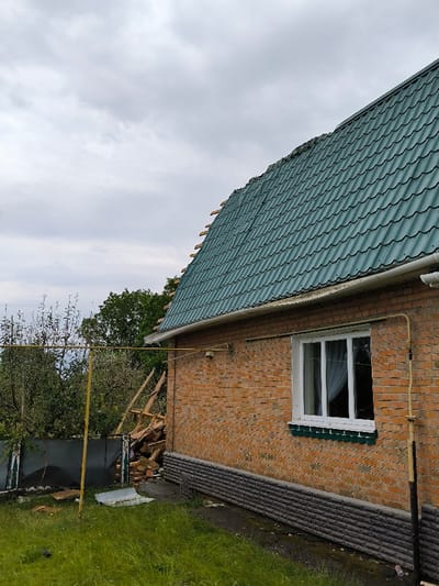 The image displays a brick building located in Pysarivka, Ukraine, under an overcast sky. The single-story structure features orange-red brick walls and a steeply pitched roof covered with green, tile-patterned metal sheeting. The upper left portion of the roof shows damage or incomplete installation, with exposed wooden rafters. A white-framed, multi-pane window with white curtains is visible on the brick facade, featuring a green-colored sill. A white plastic gutter runs along the lower edge of the roof. Yellow gas pipes are affixed horizontally and vertically to the brick wall. The building's foundation is clad with a dark gray, textured material. To the left of the building, a substantial accumulation of debris, consisting of wooden planks and bricks, is present behind a damaged, dark-colored fence. A tall, yellow metal pole structure stands in the foreground to the left. The ground is covered with green grass and patches of bare soil. Dense green trees and foliage are visible in the background. No individuals or actions are depicted.