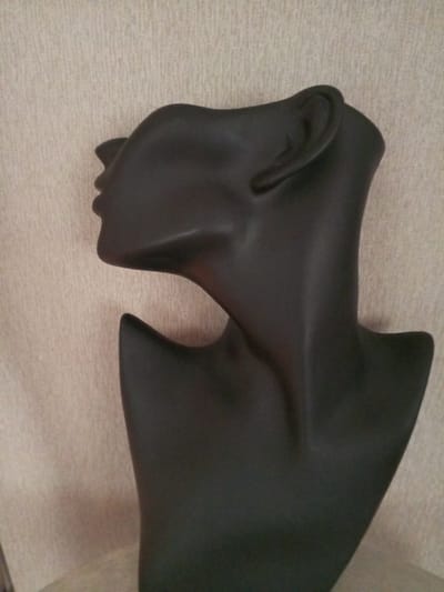 A black mannequin bust of a woman's head and neck is displayed against a textured beige background. The mannequin is positioned in profile, facing left, with its head tilted back slightly. The background appears to be a fabric wall or curtain. The lighting is subdued, casting soft shadows that highlight the contours of the mannequin's features. There are no other objects or people visible in the image. The scene is static and appears to be indoors, possibly in a retail or display setting. The location in Vilyuysk, Russia, is not determinable from the visual information provided.