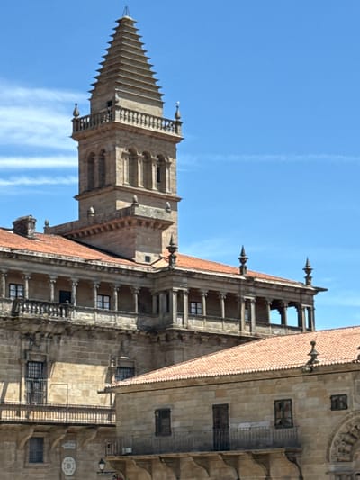 The image depicts a detailed view of the Obradoiro facade of the Santiago de Compostela Cathedral, specifically focusing on its impressive tower and surrounding architectural elements. The scene is bathed in bright sunlight, casting distinct shadows and highlighting the intricate stonework. The sky above is a clear, vibrant blue, with a few wispy white clouds present.
The central focus is a tall, ornate stone tower with a layered, pyramidal spire. This tower exhibits classical architectural features, including arched openings and a decorative balustrade. Extending from the tower, and forming a significant portion of the visible facade, is a multi-story building characterized by a loggia with classical columns and red tile roofing. This structure features numerous windows and balconies, showcasing weathered stone facades.
In the foreground, another section of the cathedral complex is visible, also with red tile roofs and stone construction. This lower building displays similar architectural styles with windows and balconies, contributing to the overall impression of a grand, historic edifice. The stonework throughout the image appears ancient and well-maintained, suggesting a place of historical and architectural significance. There are no people or animals visible in the frame. The overall environment is urban, defined by the monumental architecture of the cathedral.