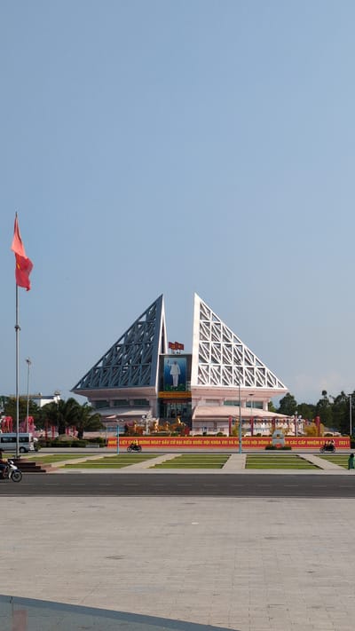 The image shows a view of a public square in the city of Phan Rang – Tháp Chàm, Vietnam, featuring a building with a distinctive architectural design. The building has two triangular structures atop, one white and the other gray, both with a lattice-like framework. A large banner with Vietnamese text is visible in front of the building. A Vietnamese flag waves on a pole to the left. The foreground is occupied by a paved plaza that extends to the right. The sky is a clear blue. Vegetation is visible along the sides of the plaza, including trees and grassy areas. A few parked scooters or motorcycles are along the roadside, and banners are visible along the building front.