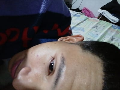 The image depicts a close-up, angled view of an individual in an indoor setting identified as Soacha ciudad, Colombia. The subject's right facial profile is prominent, featuring light to medium brown skin with minor epidermal blemishes on the forehead and cheek. Dark, short hair frames the face. The visible right ear contains a small, clear stud earring in the helix. The right eye is open, and the lips are slightly parted. A dark blue fabric, possibly a blanket or garment, with partially visible red lettering, occupies the upper left portion of the frame. The background reveals a bed covered with a light green sheet patterned with cartoon animal faces in blue, grey, and white. To the upper right, a white wall is visible, along with folded white textiles, a crumpled pink garment, and a wall outlet or switch, consistent with a bedroom environment.