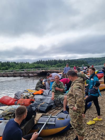 The image depicts a group of people gathered near a river in what appears to be a staging area for a rafting or boating expedition. Several inflatable rafts and associated gear, including bags and life vests, are visible on the shore and in the water. The people are dressed in casual and outdoor-appropriate clothing, some in camouflage attire. A wooden bridge or structure spans the river in the background, with a dense forest bordering the opposite bank. The sky is overcast, suggesting a cloudy or possibly rainy day. The scene conveys a sense of preparation and anticipation for an outdoor activity. While the location is stated to be the city of Вишерогорск, Russia, this is not visually verifiable from the image itself.