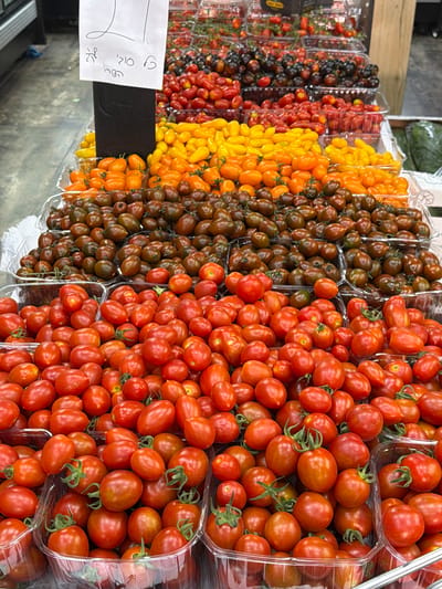 A close-up, eye-level shot shows a display of assorted tomatoes at a market stall in Ashdod, Israel. The tomatoes are packaged in clear plastic containers, stacked in tiers. The foreground is dominated by containers of small, round red tomatoes, with some cherry tomatoes mixed in. Behind these are containers of darker, plum-shaped tomatoes, and then a row of bright yellow and orange tomatoes. Further back, various other tomato varieties are visible, including red cherry tomatoes and darker, almost black tomatoes. A handwritten sign in Hebrew with the number "21" is prominently displayed on the left side of the image, with other Hebrew text below it. The background is blurred, but appears to be an indoor market setting with a concrete floor and some shelving. The lighting is bright and even, suggesting artificial light. There are no people visible in the image.