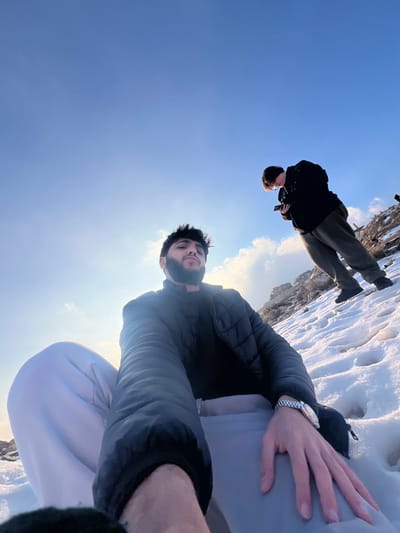 Two males are present in a snow-covered outdoor environment, specifically a hillside in Talfita, Syria, under a clear blue sky.
In the foreground, a male with dark hair and a beard, wearing a black puffer jacket, a black undershirt, and light-colored pants, is seated or crouching in the snow. His left arm is extended towards the camera, suggesting he is taking a selfie. A silver-colored watch with a metal band is visible on his left wrist. The sun is positioned behind and to the left of this individual, casting a bright glare and slight lens flare.
In the mid-ground, further up the snow-covered slope and to the right, a second male is standing, wearing a dark jacket and dark green or grey pants. He is looking down intently at a mobile phone held in both hands.
The ground consists of a layer of white snow with visible footprints and undulations. Rocky terrain and patches of bare ground are visible on the upward slope in the background. The overall lighting indicates a bright, sunny day, with long shadows implied by the low sun angle.
