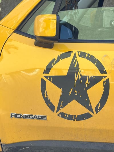 A close-up shot from above shows the hood of a yellow Jeep Renegade, featuring a large, black, distressed star enclosed in a circle. The word "RENEGADE" is displayed vertically in chrome lettering on the hood near the right edge. A portion of the driver's side window and side mirror are visible on the left side of the frame. The scene suggests the vehicle is parked outdoors. While the location is Saguramo, Georgia, specific details tying it to that city are not discernible from the image alone.