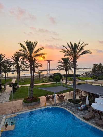 The image depicts a serene beachside resort at sunset in the Hlorakas Community, Cyprus. In the foreground, a bright blue swimming pool is visible with two children playing near the steps, one wearing a yellow float and another with a snorkel mask. The pool area is surrounded by a wooden deck and a stone patio with lounge chairs, some occupied by guests. Several large white umbrellas provide shade.
Beyond the pool, a well-maintained lawn is dotted with palm trees, leading towards the beach and the sea. In the mid-ground, outdoor seating areas with tables and chairs are set up under grey canopies, some with patrons enjoying the evening. A pathway made of stone tiles winds through the grass.
The background showcases the coastline with gentle waves lapping against a rocky shore. The sun is setting over the horizon, casting a warm, orange and pink glow across the sky and reflecting on the water. A distinctive modern structure with a large, circular top element stands near the sea. The scene conveys a relaxed and picturesque holiday atmosphere.