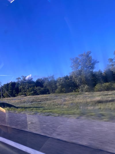 The image shows a bright blue sky with a few wispy clouds. On the right side of the frame, there is a road with white markings, suggesting a highway or paved road. Adjacent to the road, there is a grassy area and a dense line of trees. Some of the trees are tall and full, while others appear more sparse. In the distance, a plume of white smoke or steam rises from behind the trees, partially obscured by foliage. A faint contrail from an aircraft is visible in the sky, stretching across the blue expanse. The overall impression is of a sunny day, possibly late morning or afternoon, in a rural or semi-rural setting. The image appears to be taken from a moving vehicle due to the motion blur on the road and the perspective. There are no visible people or discernible text in the image.